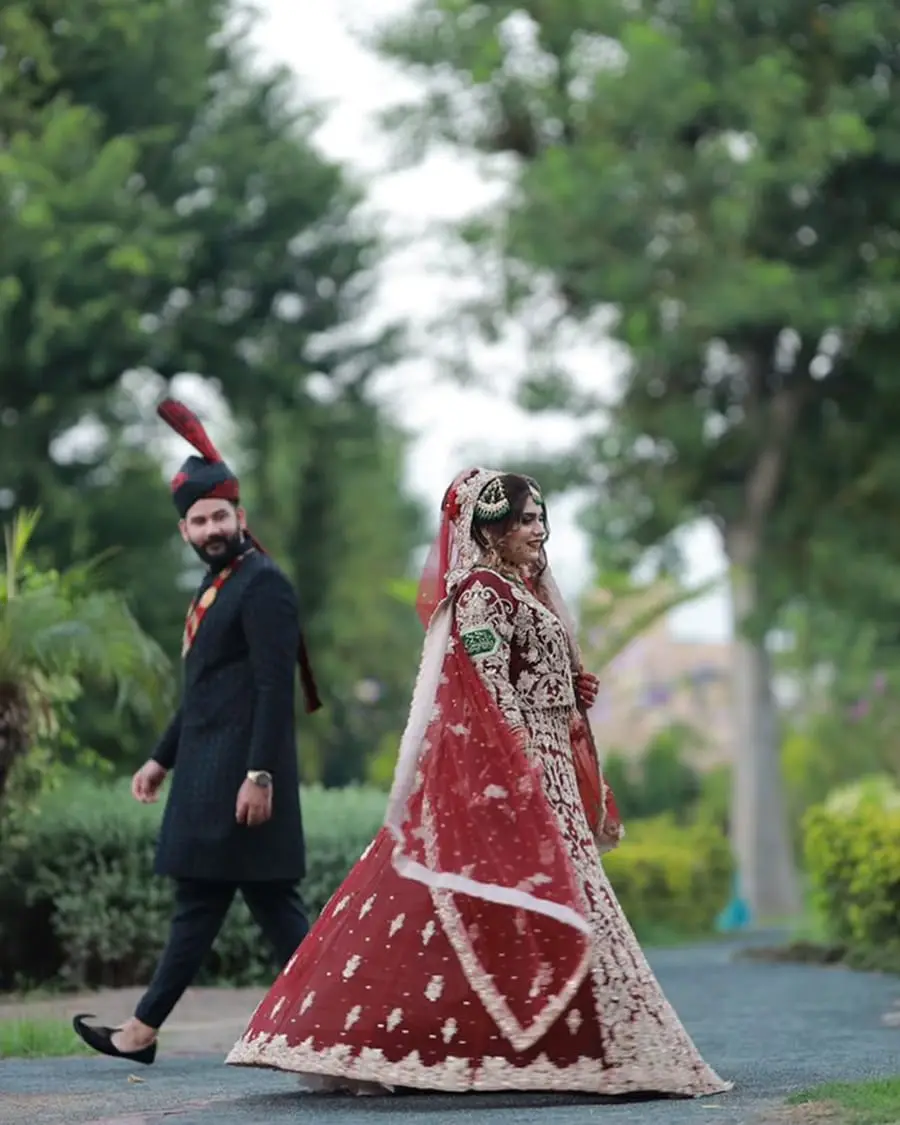 Bride in a flowing red lehenga walking ahead of the groom in a garden